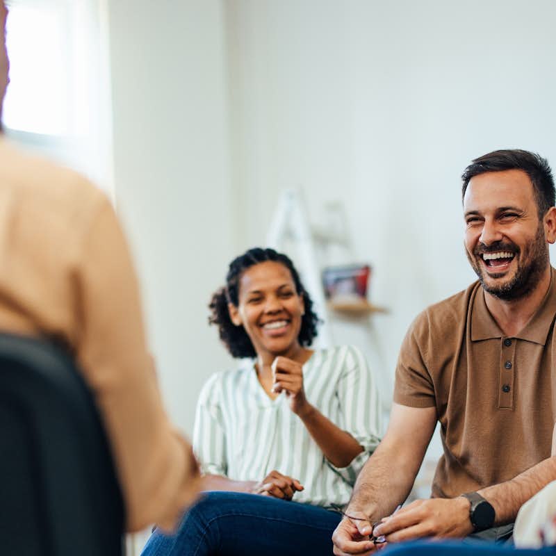 Focus on the smiling man, talking with people of all ages, during the group therapy.