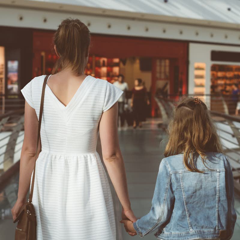 A woman in a white dress holds the hand of a little girl wearing a denim jacket.